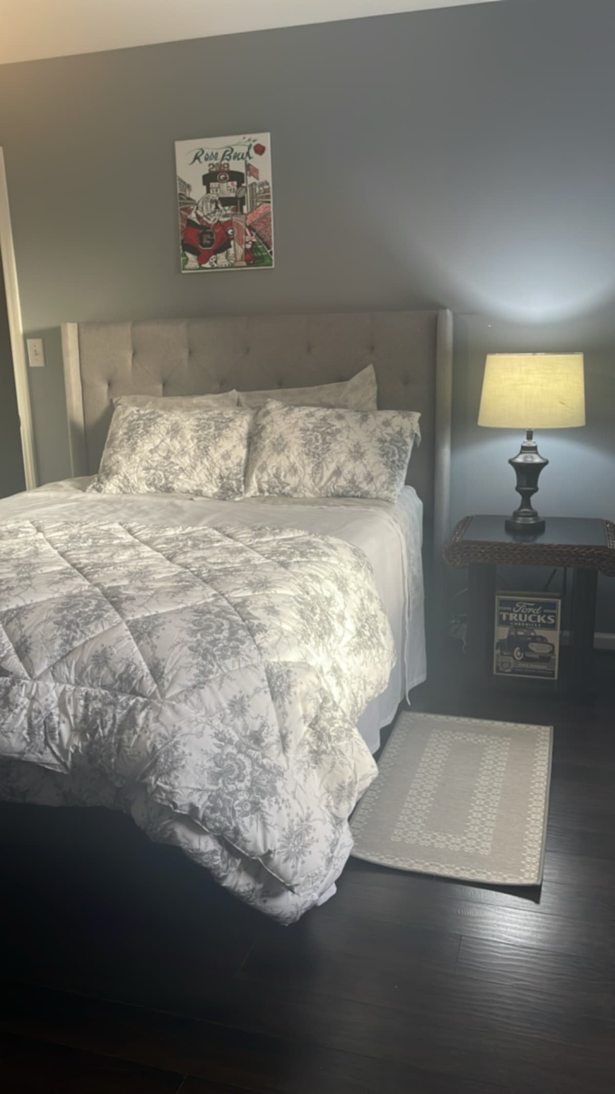 A cozy bedroom features a plush bed with elegant bedding and multiple decorative pillows. Beside the bed, a classic lamp provides soft lighting on a nightstand. A textured rug is placed at the foot of the bed, complementing the dark wooden flooring.