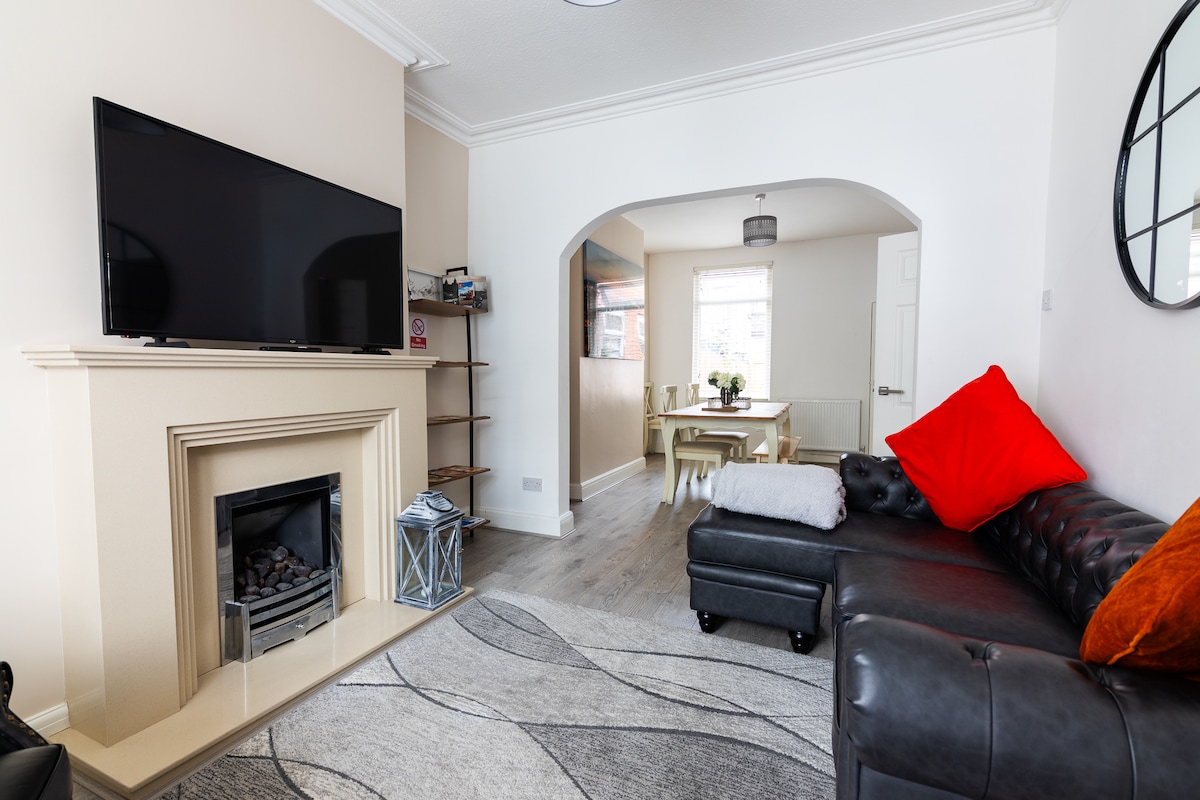 A modern living area features a sleek black television mounted above a decorative fireplace. A dark leather sectional sofa adorned with a vibrant red cushion offers comfortable seating. Light pours in from a nearby window, illuminating a dining area with a simple table and chairs in the background.