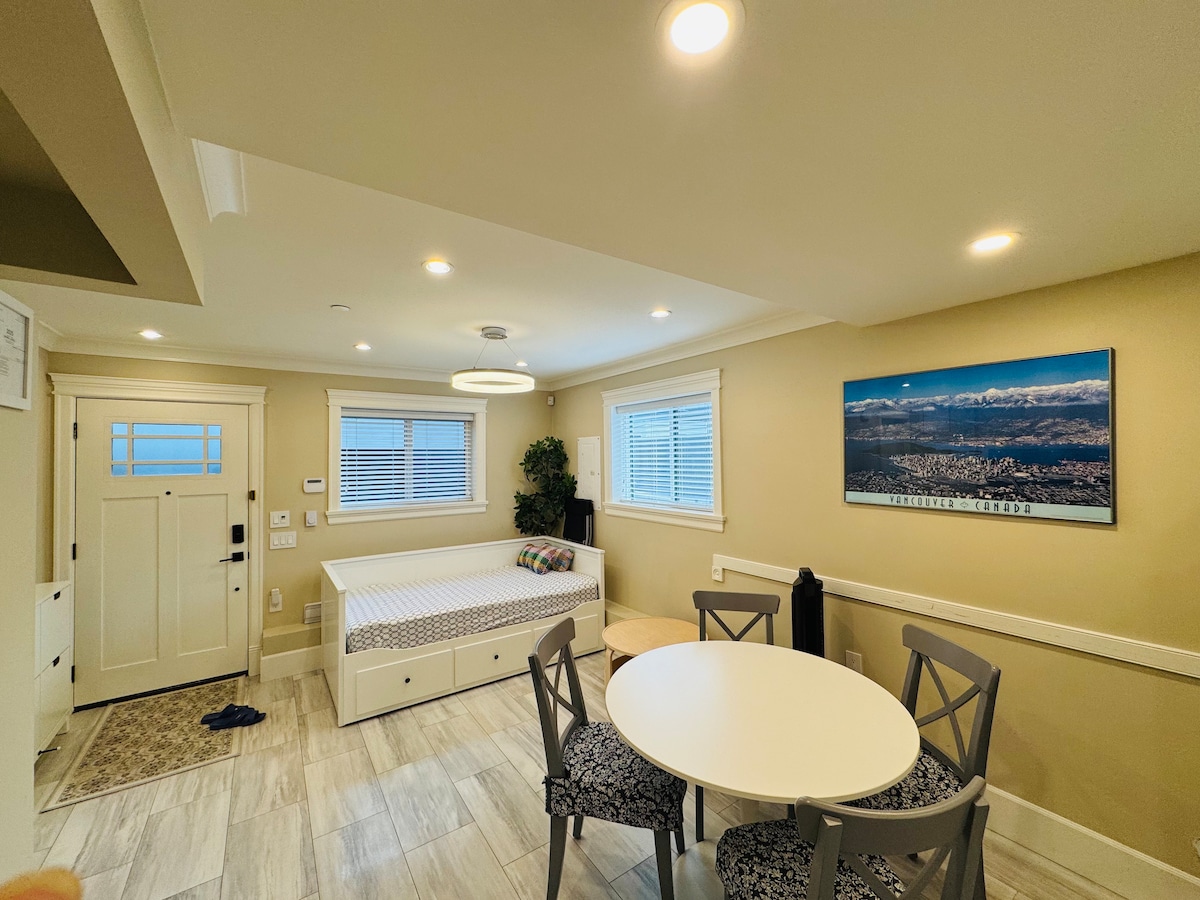 A bright and airy living space features a multi-functional bed with built-in storage beneath, surrounded by large windows allowing natural light. A round dining table with four chairs is positioned near the entrance. A scenic landscape photo adorns the wall, enhancing the room's ambiance.
