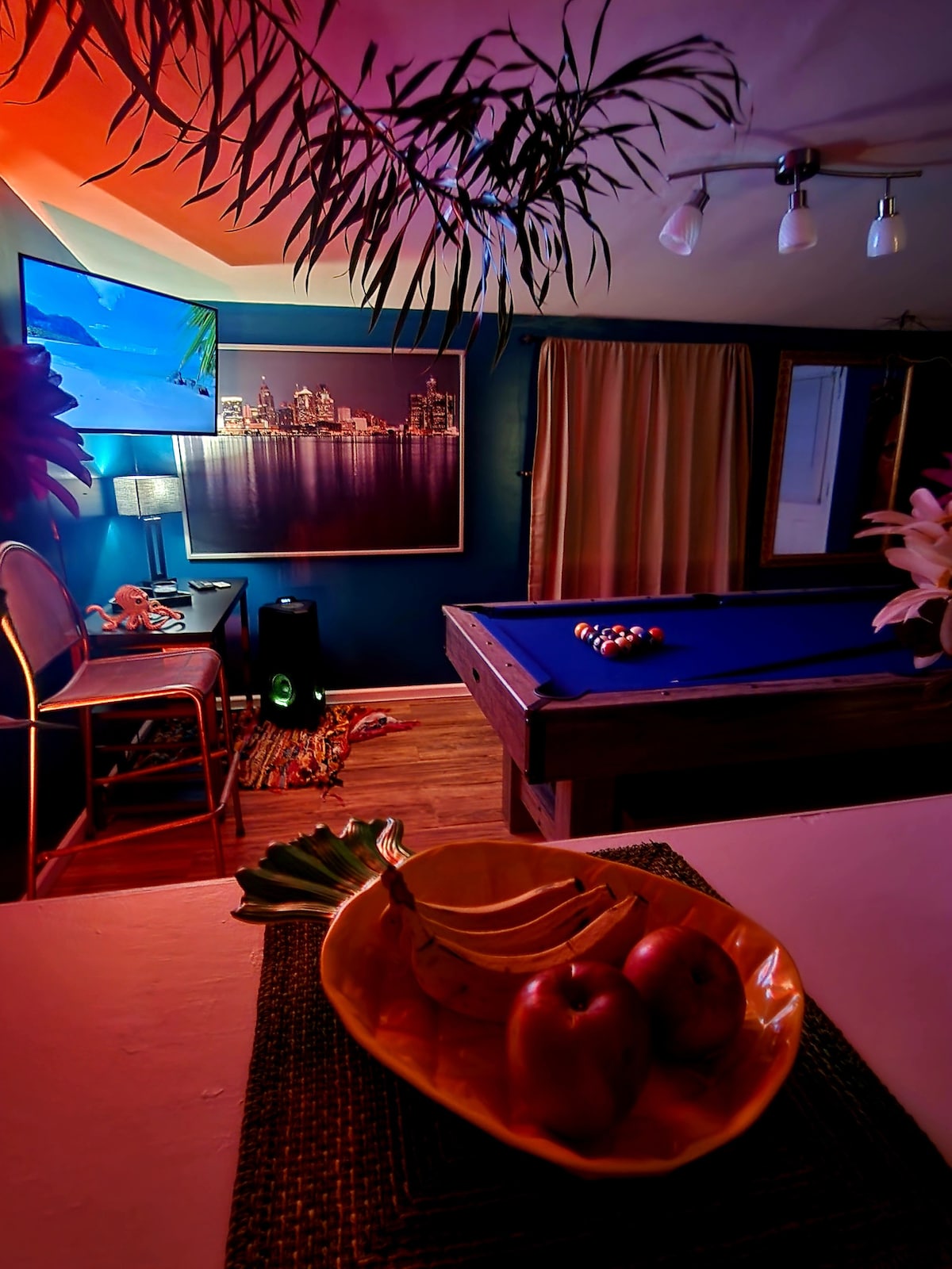 An inviting theater and recreation space features a pool table with colorful balls arranged for play. A television hangs on the wall, and soft ambient lighting creates a relaxing atmosphere. A decorative bowl with apples and bananas is placed on a nearby table, adding a touch of color.