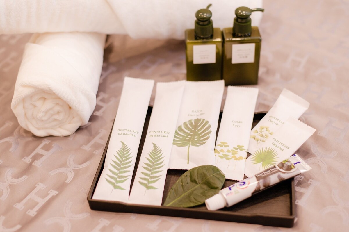 A neatly arranged set of personal care items is displayed on a dark tray. This includes dental kits, a tube of toothpaste, and two green bottles of toiletries. A rolled white towel provides a spa-like feel, enhancing the overall minimalist presentation.