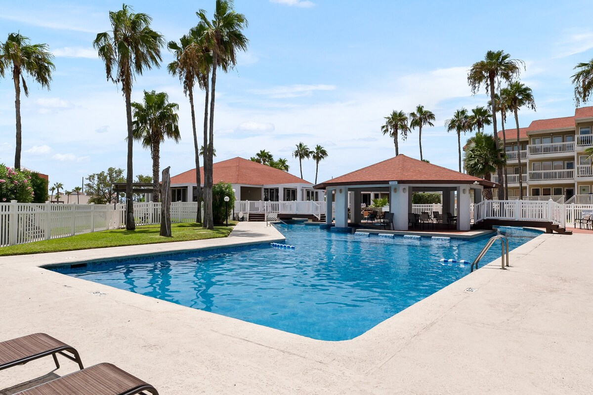 A large swimming pool is surrounded by palm trees, with lounge chairs positioned nearby. Two covered pavilion areas are visible, providing shaded spots for relaxation. The well-maintained pool area features clear blue water and a light-colored deck, set against a backdrop of lush landscaping.