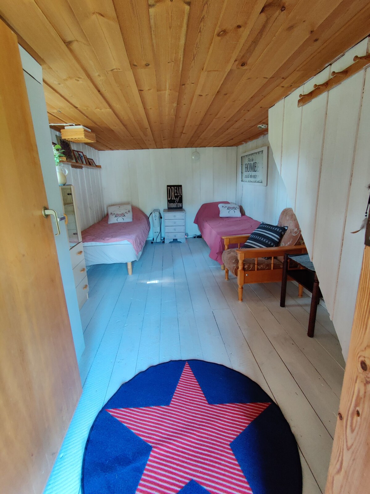 A cozy interior is presented, featuring two spacious beds with light bedding arranged in parallel. A small nightstand with decorative items can be seen beside one bed. A wooden armchair adds a touch of seating, complemented by a circular rug with a star pattern on the floor.