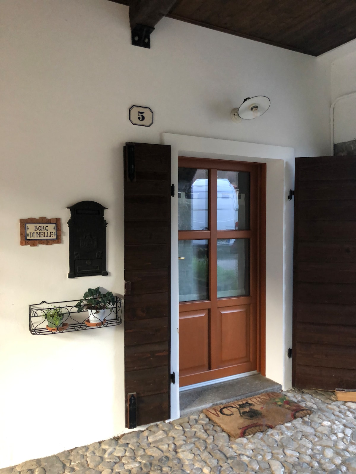 The entrance features a traditional wooden door with large glass panes, framed by dark wooden shutters. A small shelf with potted plants is mounted beside the door, contributing to a welcoming appearance. The entryway is lined with smooth stones, enhancing the charming entrance.