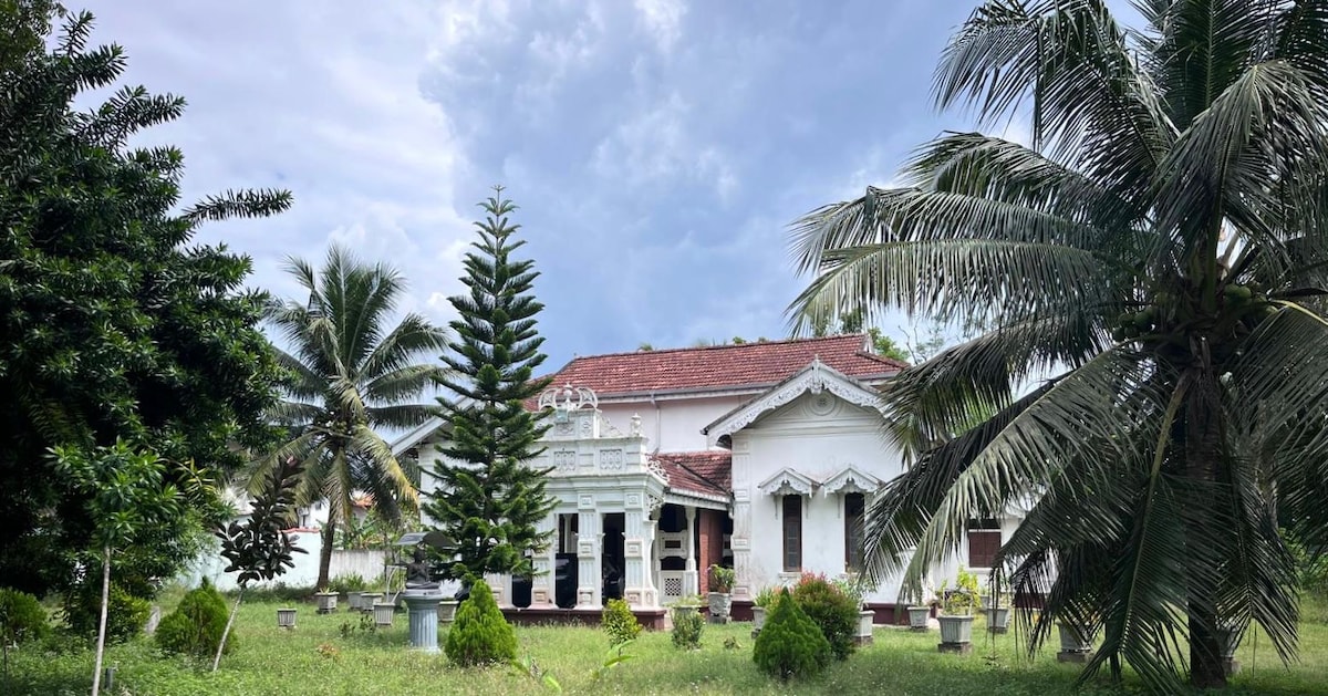 A colonial-era house is set in a lush green garden surrounded by tall palm trees. The structure features intricate architectural details, including a decorative porch with columns. The sky above is partly cloudy, creating a tranquil atmosphere in the outdoor space.
