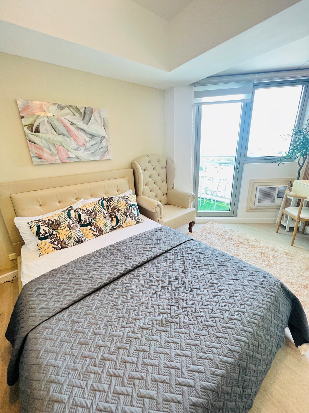 A light-filled bedroom features a spacious bed with a patterned headboard and a decorative throw blanket. An upholstered armchair is positioned nearby, alongside a small dining table. A large window offers a view, while light-colored walls and a soft rug contribute to the room's modern decor.