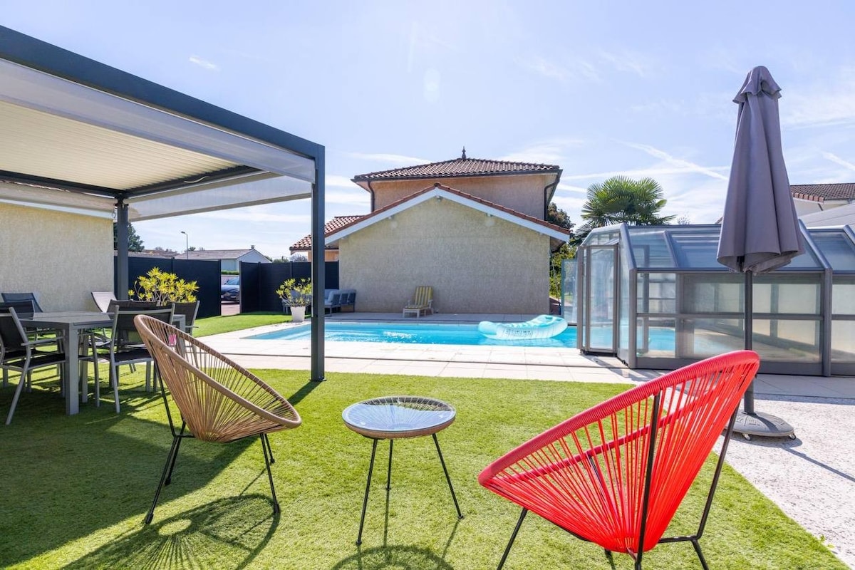 An outdoor area is present, featuring seating arrangements with colorful chairs and a small table on manicured grass. A pool glimmers in the background under a clear sky, with a covered terrace nearby providing shade. A parasol stands at the edge, completing the inviting space.