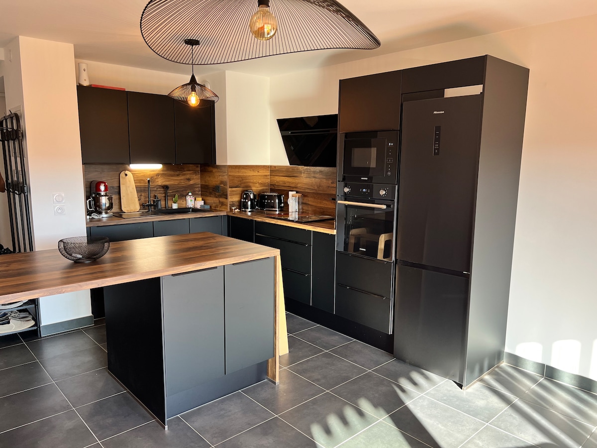 A modern kitchen features sleek black cabinetry with wood accents and stainless steel appliances. A central island with a wooden countertop stands in the middle, while pendant lighting adds gentle illumination. The space is complemented by dark tile flooring, providing a clean and contemporary appearance.