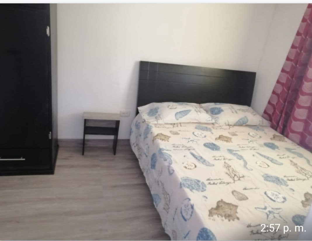 A simple bedroom features a spacious bed adorned with a patterned bedspread, a small nightstand is positioned beside it. A dark wardrobe stands against the wall, while light-colored flooring enhances the room's openness.