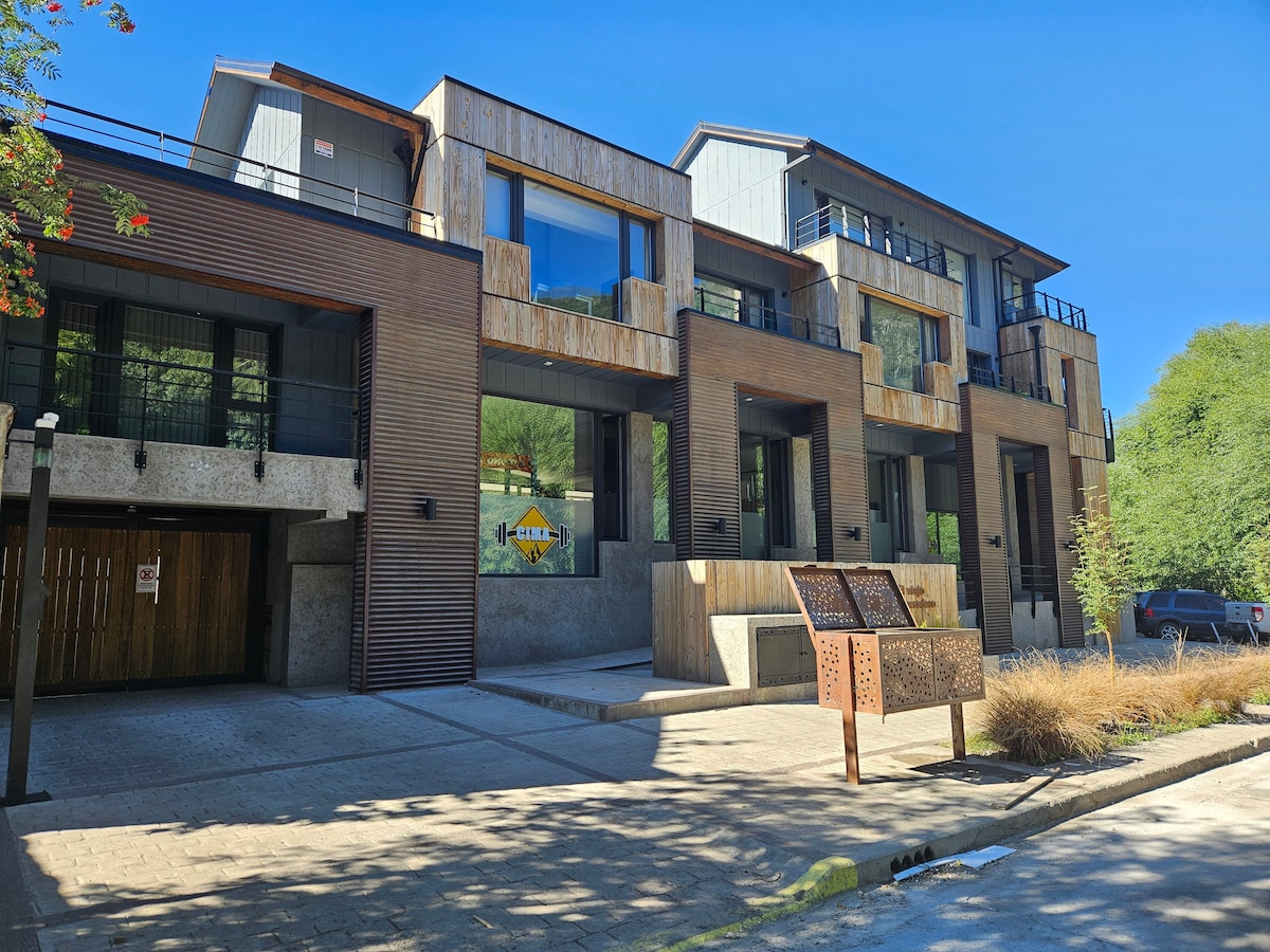 The modern building features a mix of wood and metal facades, along with large windows that allow natural light to enter. Balconies extend from the upper floors, creating outdoor spaces. A paved entrance is lined with greenery and decorative elements, enhancing the overall aesthetic.