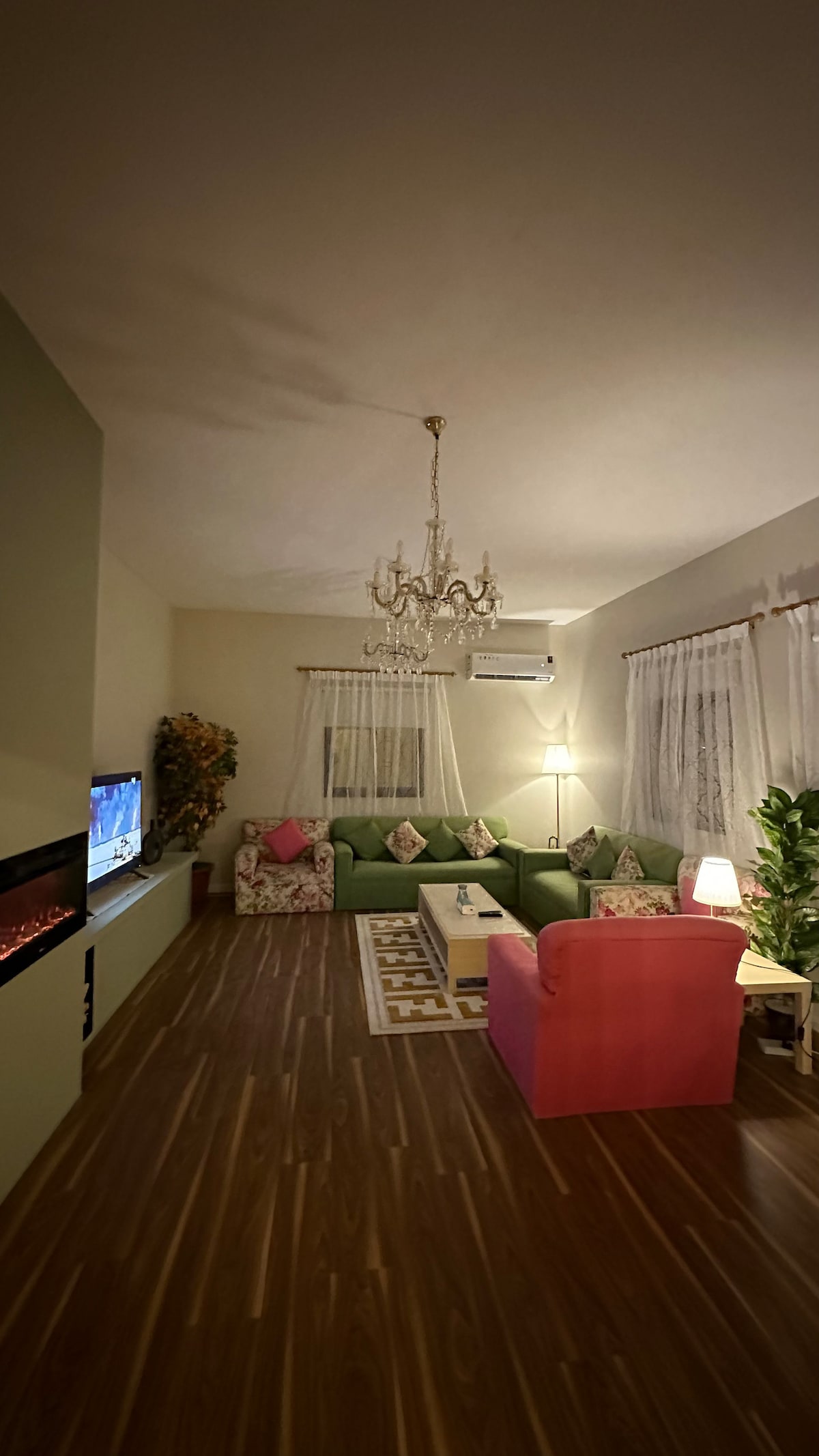A lounge area is presented, featuring a green sectional sofa adorned with patterned cushions, complemented by a pink chair. Soft lighting is provided by a modern chandelier, while an electric fireplace is positioned against the wall, offering a cozy focal point.