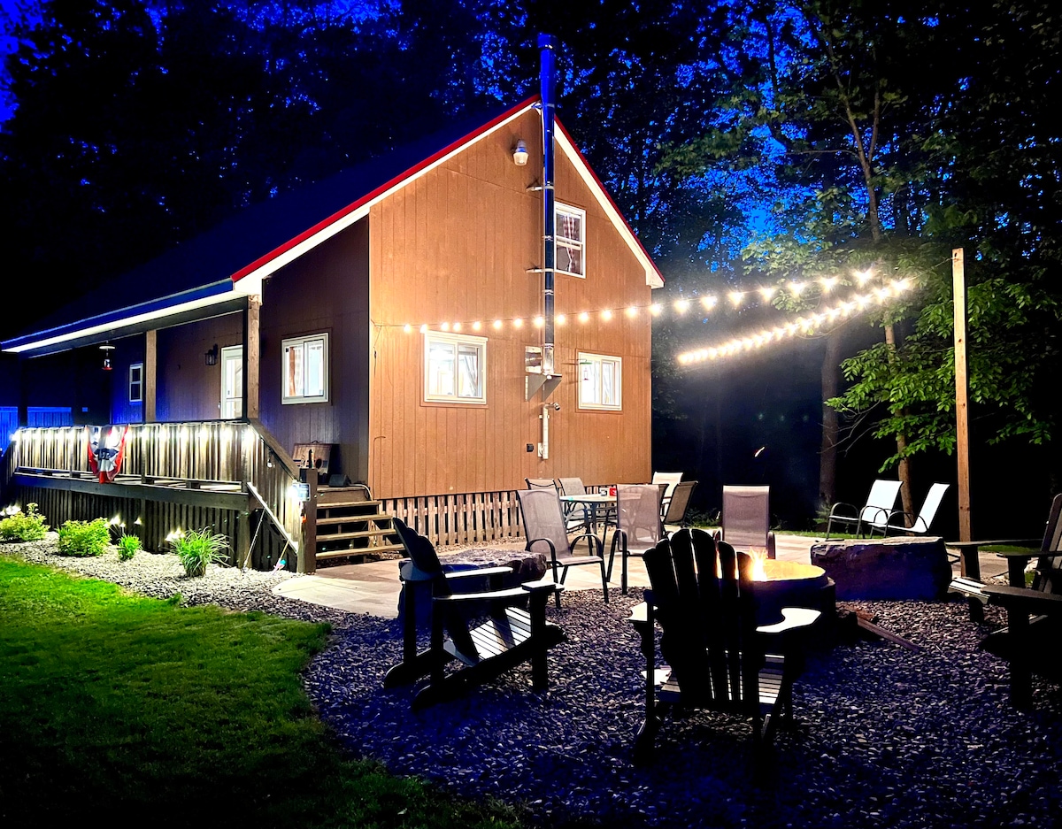 The cabin exterior is softly illuminated by string lights at dusk, showcasing the rustic brown siding and inviting covered porch. A firepit area is surrounded by several Adirondack chairs, with landscaping visible around the gravel patio, creating a cozy outdoor retreat.
