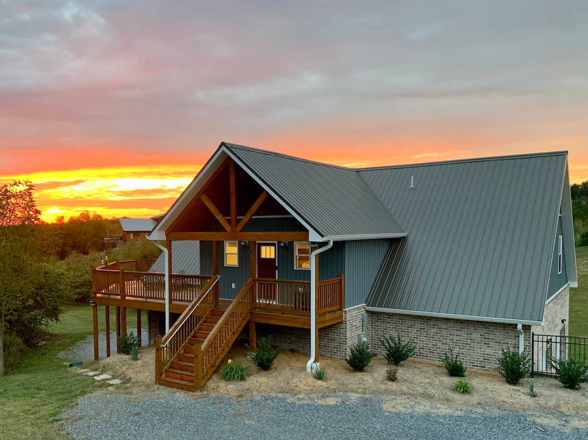 A modern guest house with a stunning sunset backdrop, featuring a peaked roof and a large front porch. The entrance is flanked by wooden railings, and the surrounding landscape includes a gravel pathway leading to the deck and landscaped shrubs.