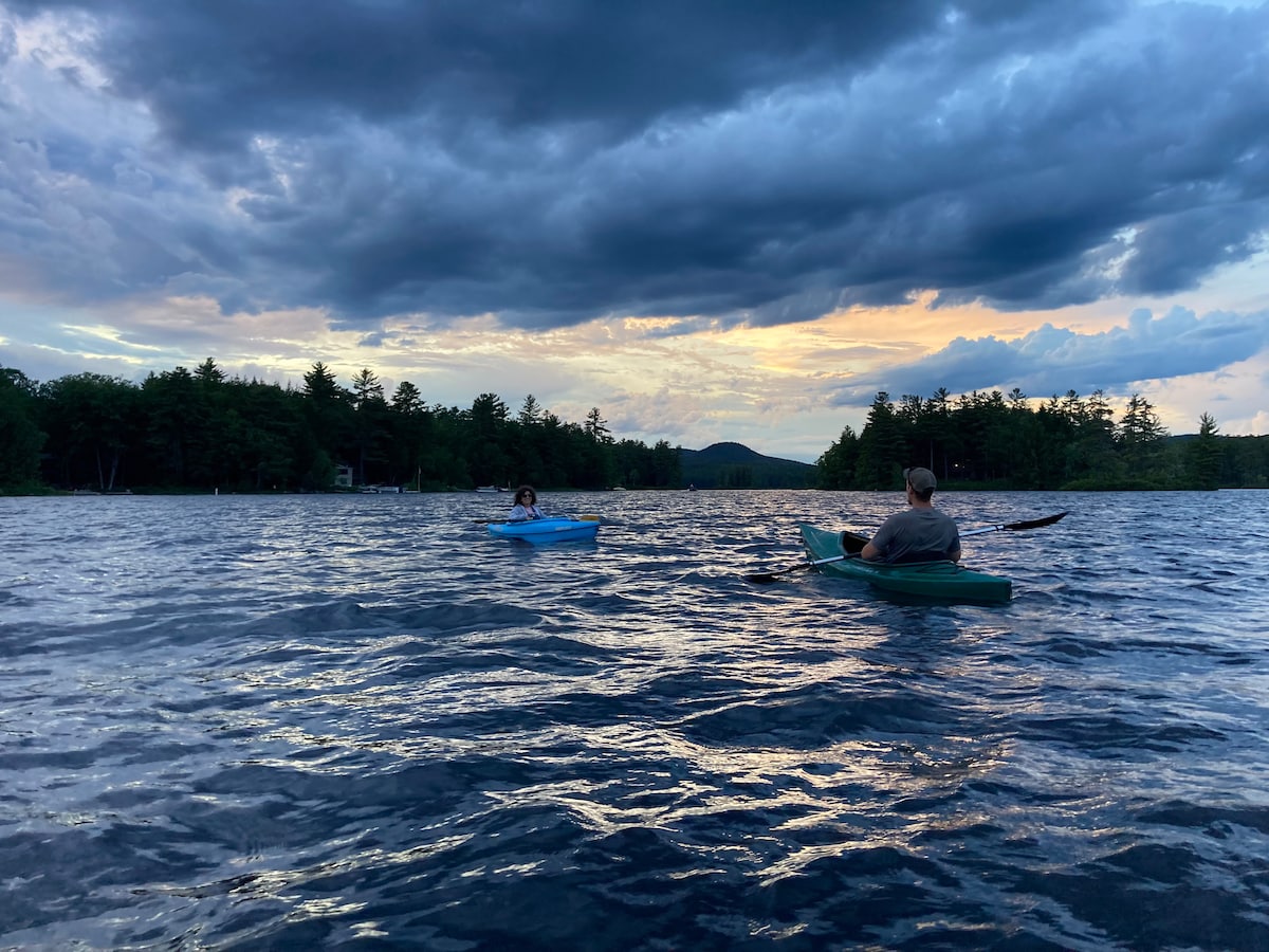Two individuals are peacefully paddling in kayaks on a tranquil lake. A backdrop of rolling clouds and a colorful sunset reflect upon the water, surrounded by lush trees and distant mountains, creating a serene outdoor setting.