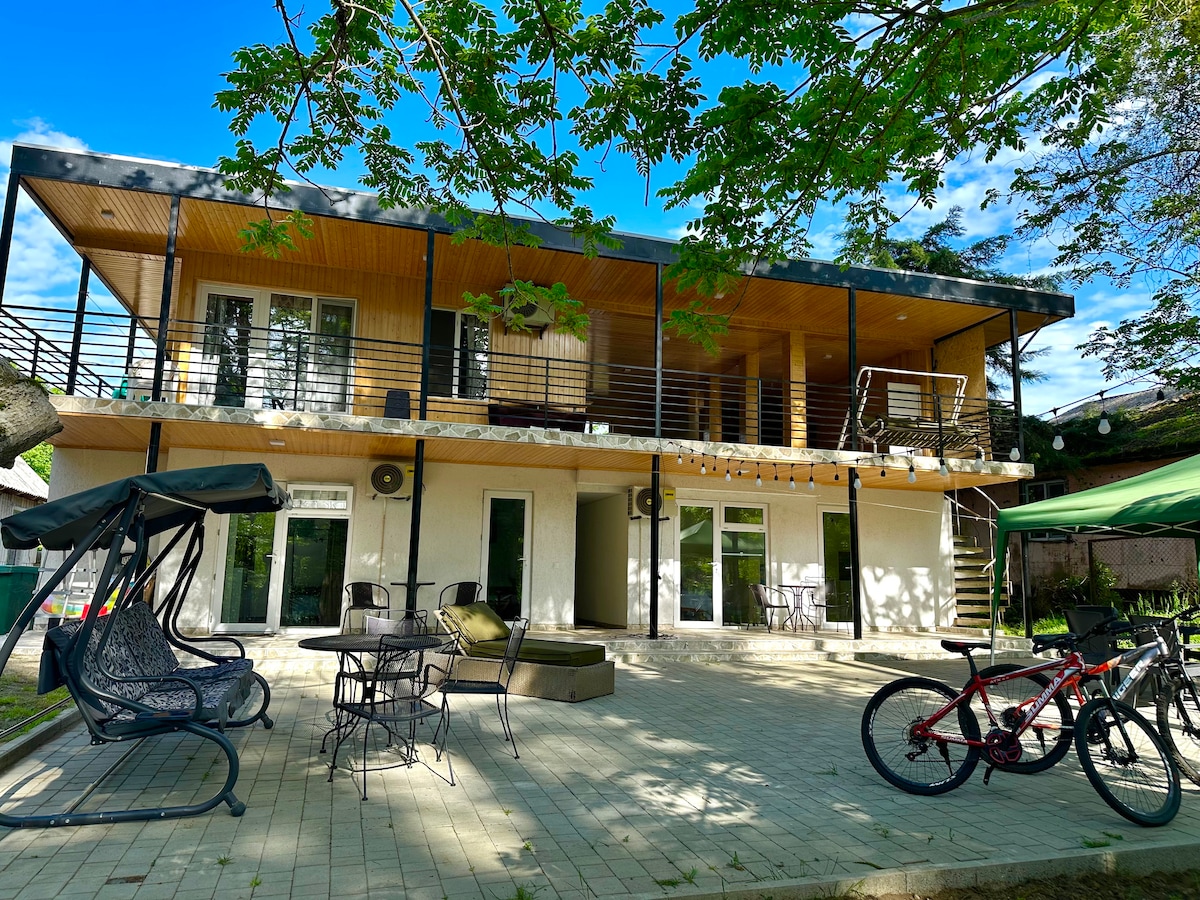 The exterior of a two-story building is showcased, featuring a spacious patio with tables and seating. A swing and bicycles are visible, surrounded by greenery. Large glass doors provide access to individual rooms, and the balcony offers a view of the surrounding area.