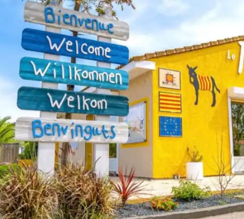 A colorful welcome sign displayed prominently features greetings in multiple languages, including French, English, German, Dutch, and Spanish. The vibrant yellow building serves as a backdrop, adorned with playful artwork, including a donkey, enhancing the friendly atmosphere of the site.