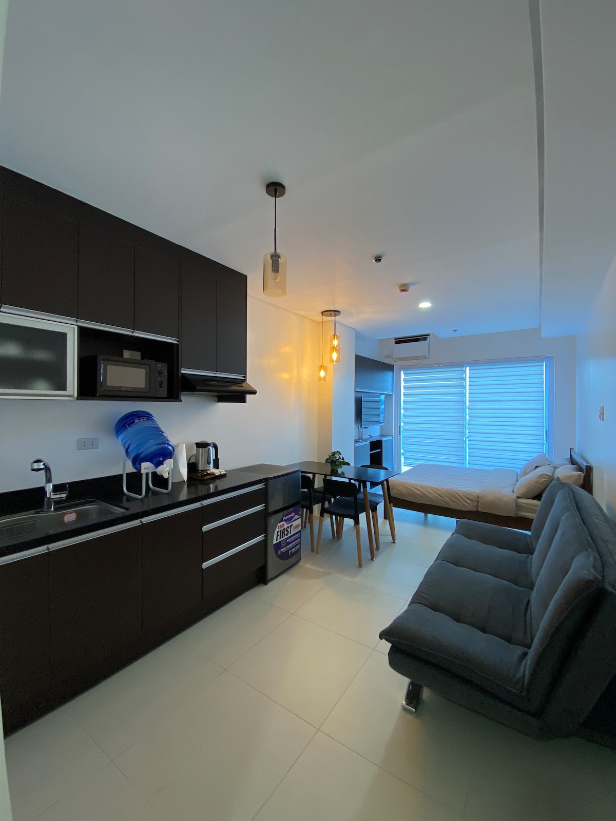A compact kitchen area features dark cabinetry with modern appliances including a microwave and coffee maker. A dining table with four chairs is positioned nearby, with a comfortable sofa and a spacious bed in the background. Natural light filters through the horizontal blinds.