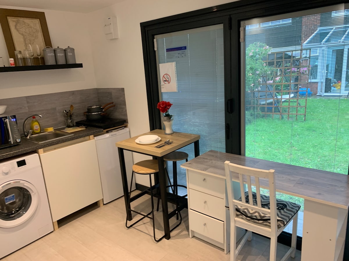 The compact kitchen includes a washing machine, a stove, and an array of cooking utensils. A table for two is positioned next to a sliding glass door that leads to a private garden, offering natural light and outdoor views.