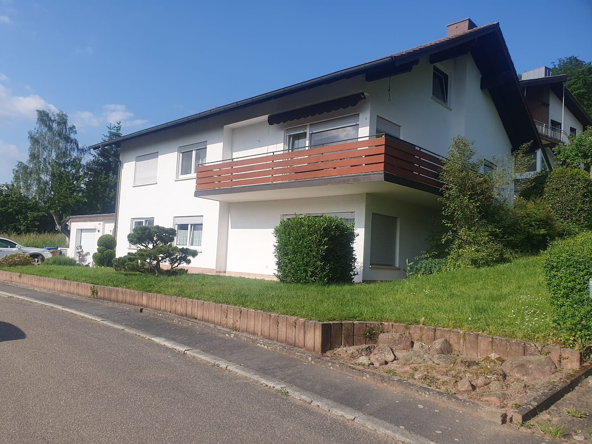 A two-story white house is set against a clear blue sky. The front yard features well-maintained greenery, including a small bush and a patch of grass. A balcony with wooden railings extends from the upper level, providing an outdoor space for relaxation.