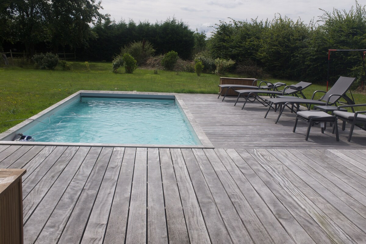 A rectangular heated pool is surrounded by a wooden deck. Several gray lounge chairs are positioned nearby, inviting relaxation. Lush greenery is visible in the background, creating a peaceful setting.