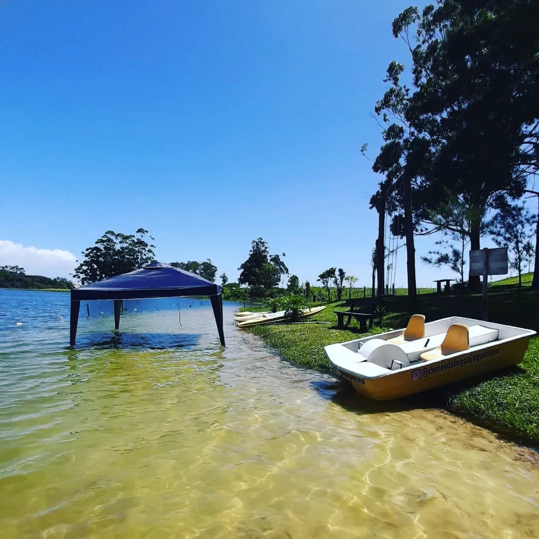 A calm lakeside scene features a small yellow boat anchored at the shore. A shaded tent sits on the grassy bank, providing shelter from the sun, while trees line the background under a clear blue sky.