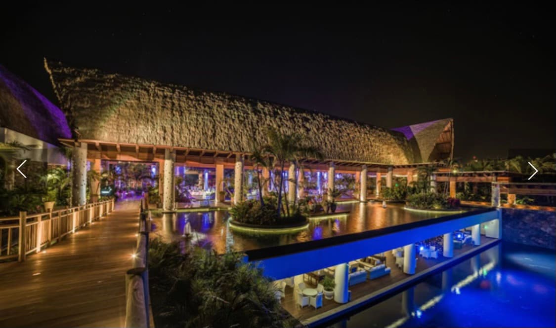 An expansive resort structure features a thatched roof design illuminated by soft purple lighting at night. Surrounding palm trees and water reflect the vibrant hues, creating a serene ambiance. Pathways lead to inviting seating areas, enhancing the overall inviting atmosphere of the resort.