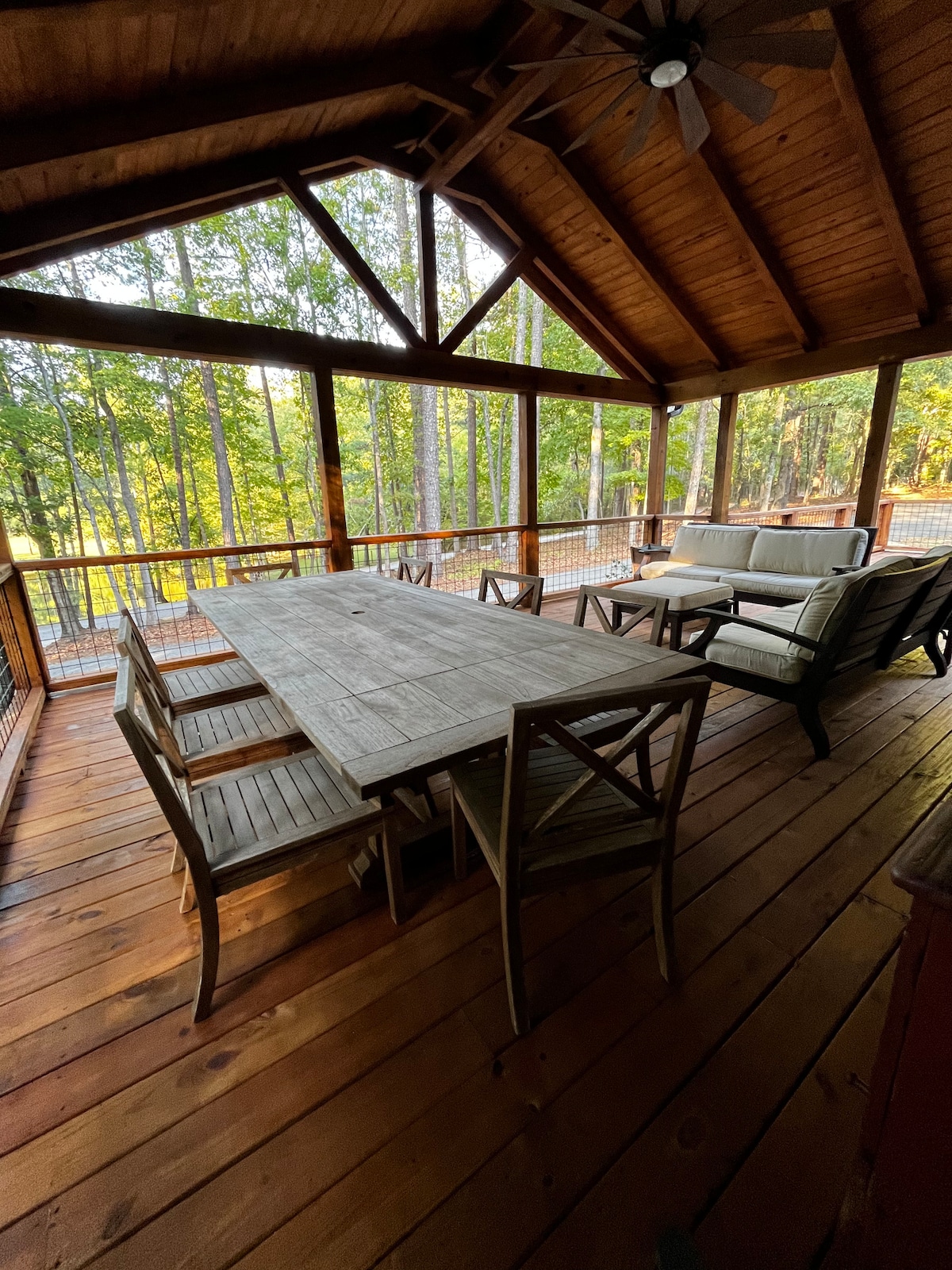 The outdoor deck features a large wooden dining table surrounded by six chairs, providing ample seating. Two outdoor couches offer a comfortable area for relaxation. The deck is framed by trees, creating a serene atmosphere, and the wooden ceiling adds a rustic touch.