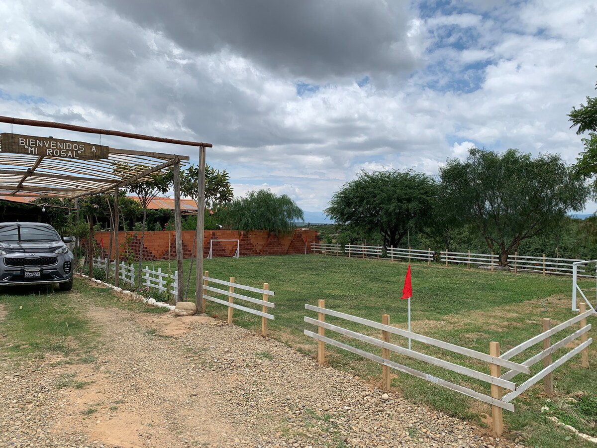 A spacious outdoor area is surrounded by greenery and wooden fencing. A neatly maintained lawn is visible, with a small red flag placed in one corner. The structure features a welcoming sign, and a vehicle is parked nearby, integrating into the peaceful, natural setting.