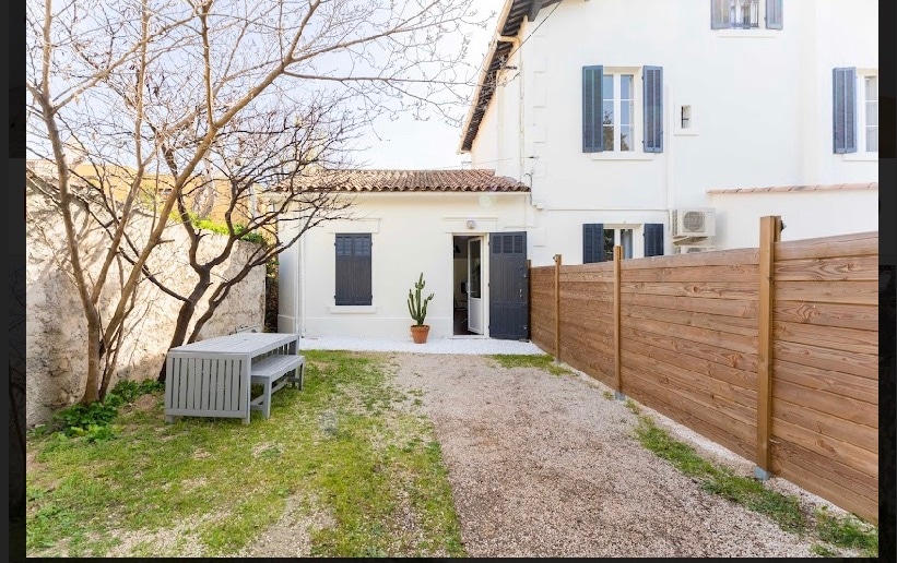 An outdoor space is featured, showcasing a wooden fence and a gravel pathway leading to a small garden area. A minimalist table with benches is positioned under the sunlight, surrounded by greenery and a potted plant. The entrance to the house is visible in the background.