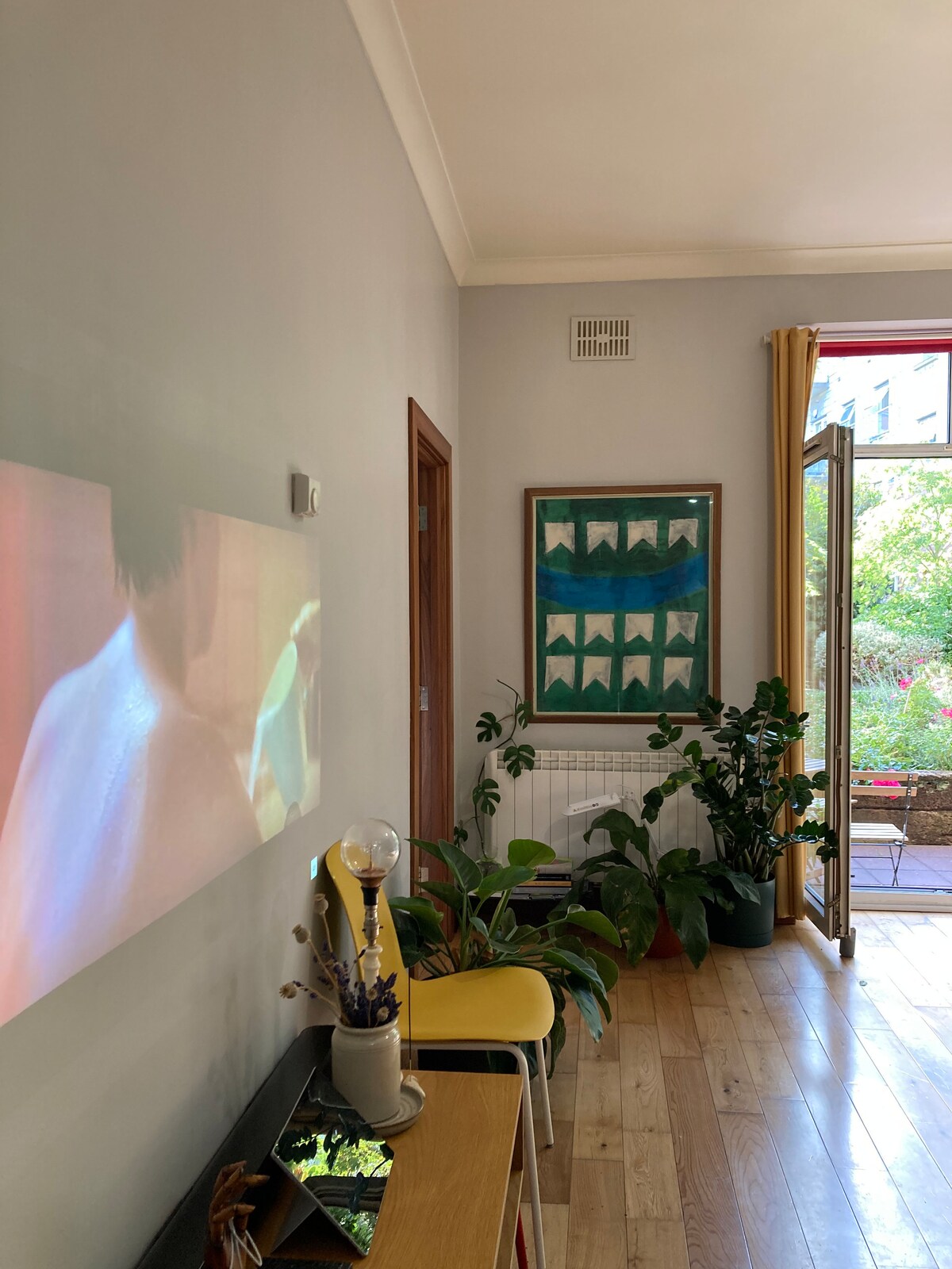 A cozy living area is highlighted by a wall-mounted projector displaying a film. Potted plants add greenery to the space, while a wooden table holds decorative items. A door opens to a courtyard filled with natural light, contributing to a welcoming atmosphere.