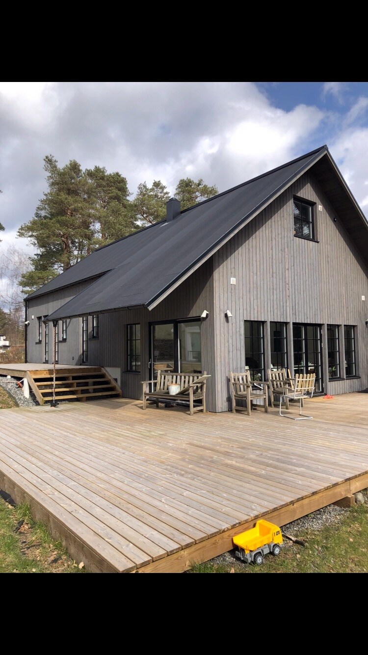 A modern wooden villa features a large deck with multiple seating areas. The façade showcases sleek gray siding and large windows that allow natural light. Surrounding trees provide a natural setting, enhancing the outdoor space that invites relaxation.
