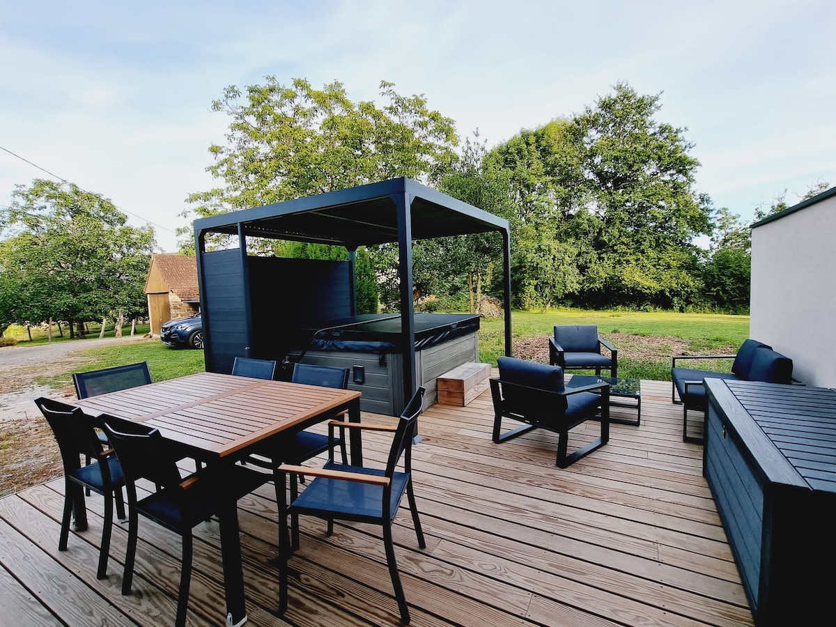 An outdoor wooden terrace features a dining table surrounded by black chairs, alongside a lounge area with comfortable seating. A covered spa is positioned in the background, with greenery and trees visible beyond the terrace.