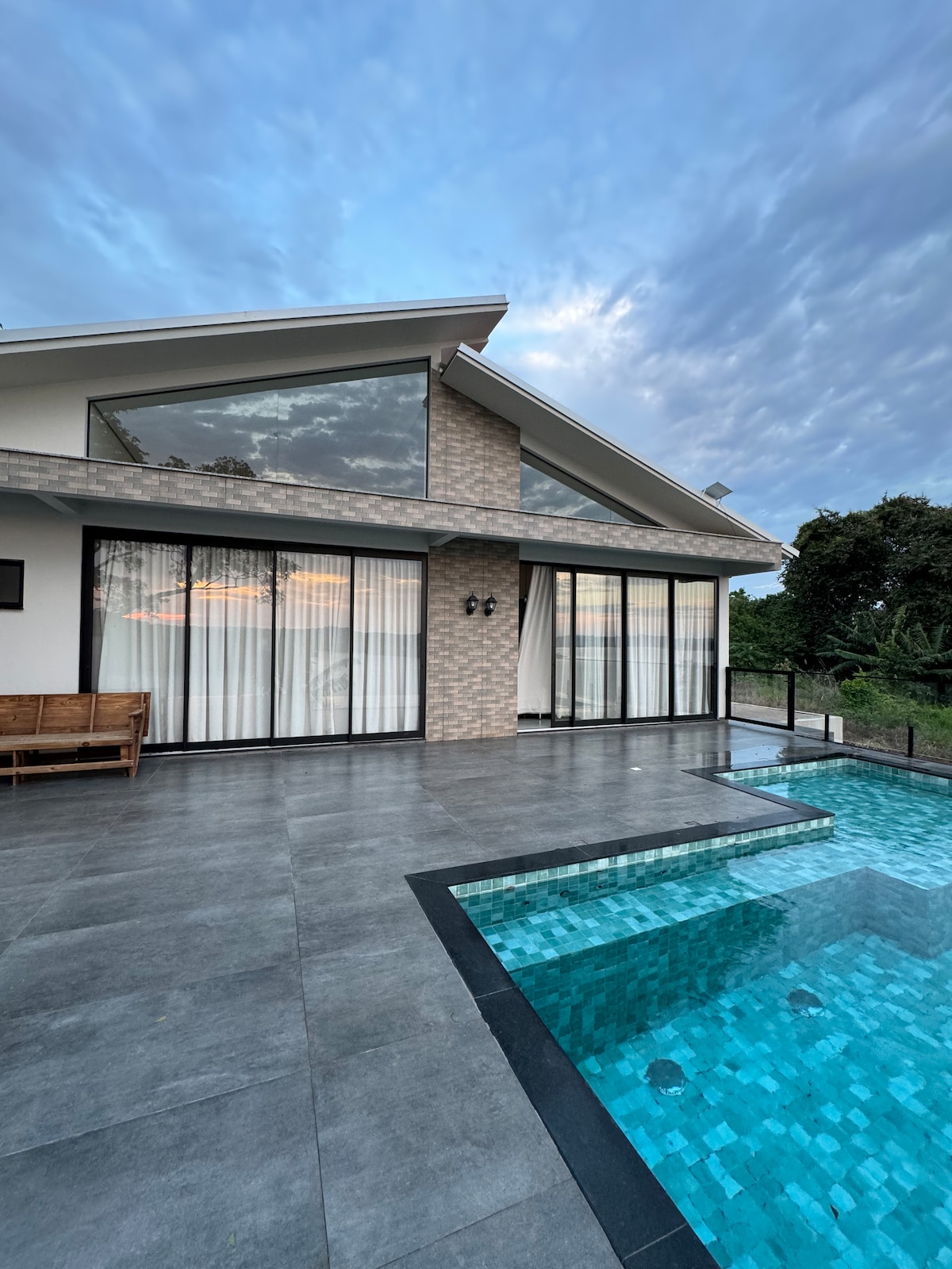 The exterior of the house is shown with large glass doors leading to a patio area. A tranquil pool with a mosaic design sits alongside the patio, reflecting the sky above. Lush greenery surrounds the property, contributing to a serene atmosphere.