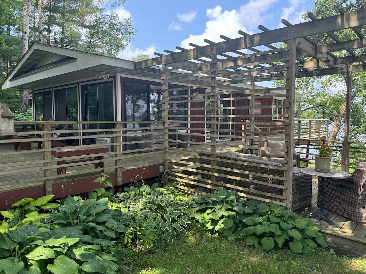 A rustic cabin exterior is framed by lush greenery and a wooden trellis. Comfortable seating arrangements, including wicker chairs, are present on a spacious deck, inviting relaxation. Large glass doors reveal the interior and provide a connection to nature and the tranquil setting by the lake.