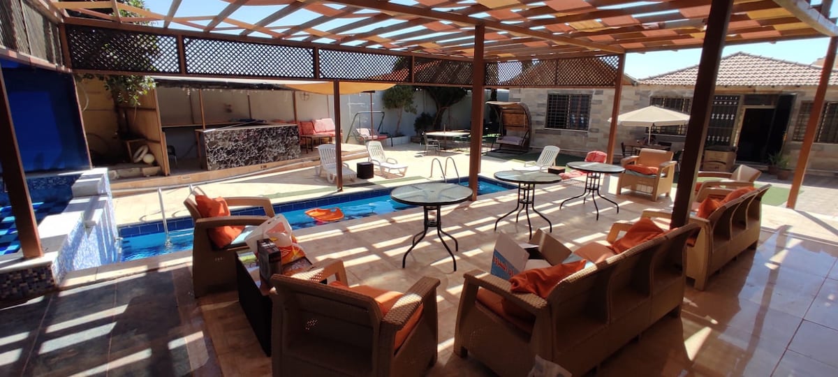 A spacious outdoor area includes a swimming pool and several seating arrangements. Two tables are positioned amidst comfortable chairs, offering ample space for relaxation. A shaded pergola provides coverage, while an outdoor bar adds functionality to the space.