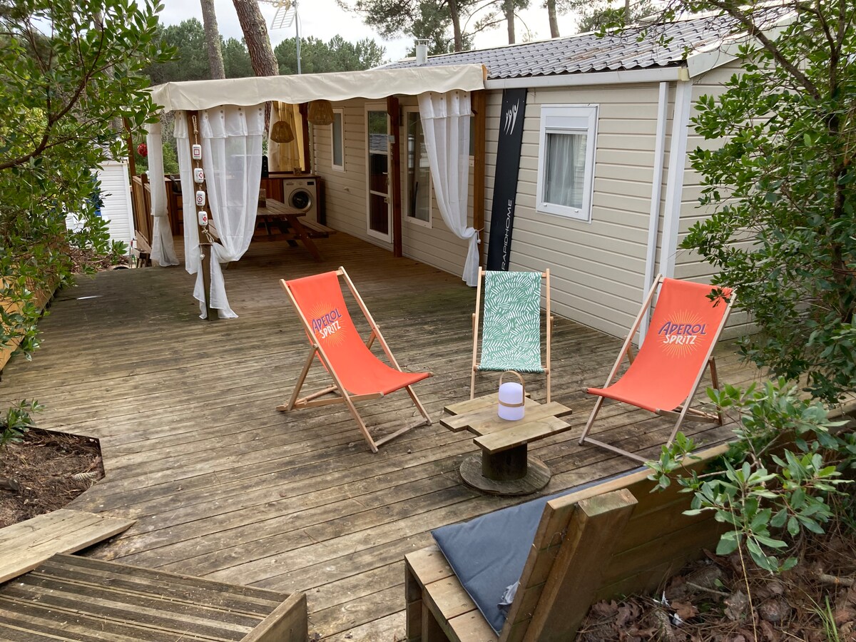 A wooden deck area features two colorful lounge chairs and a small table, surrounded by greenery. The entrance to a mobile home is visible through a canopy adorned with white curtains, creating a relaxed outdoor seating space.