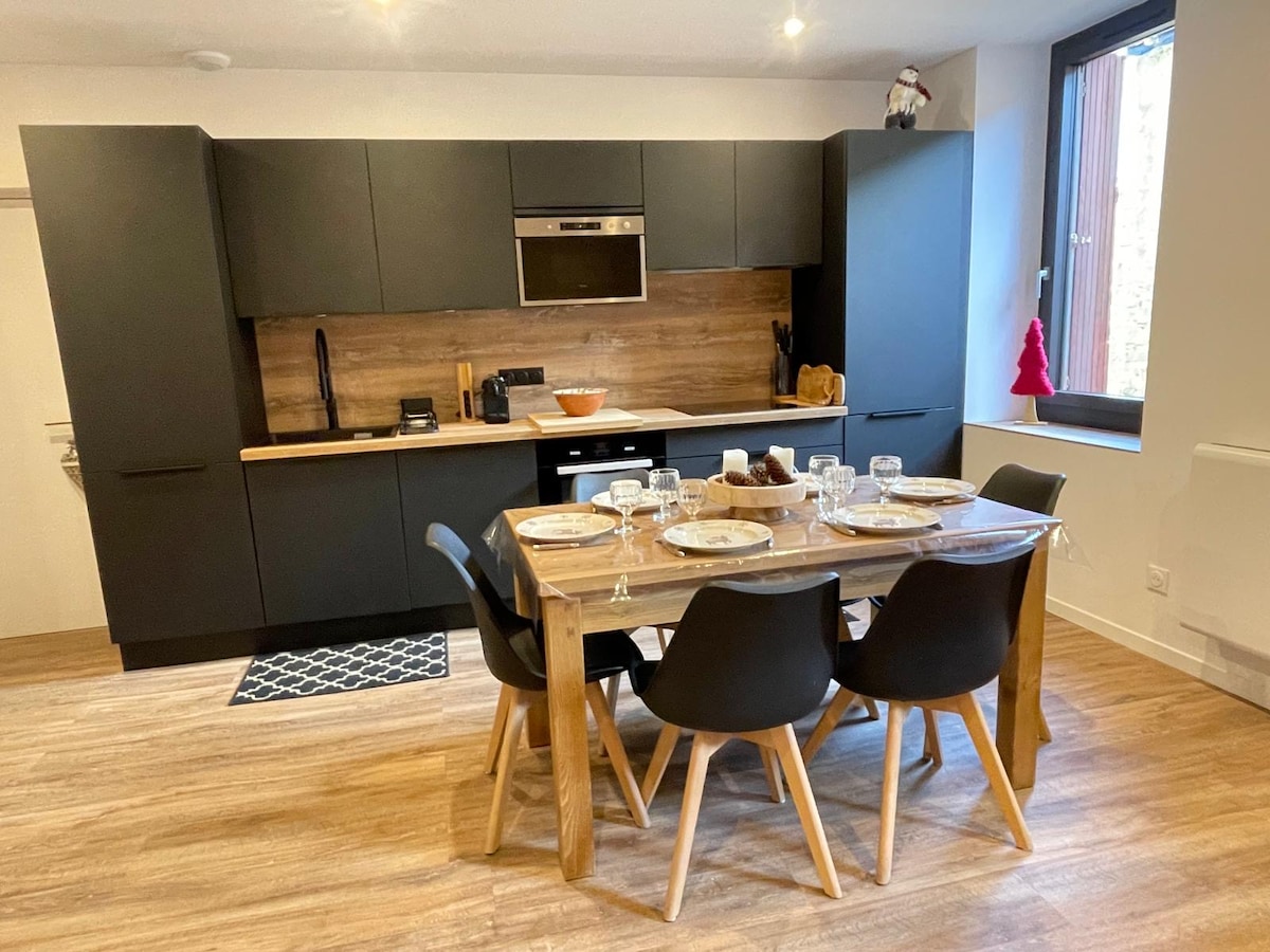 An open kitchen features sleek black cabinetry, complemented by wooden accents. A wooden dining table, seating six, is set with plates and glassware. Large windows allow natural light to fill the space, enhancing the modern and functional design.