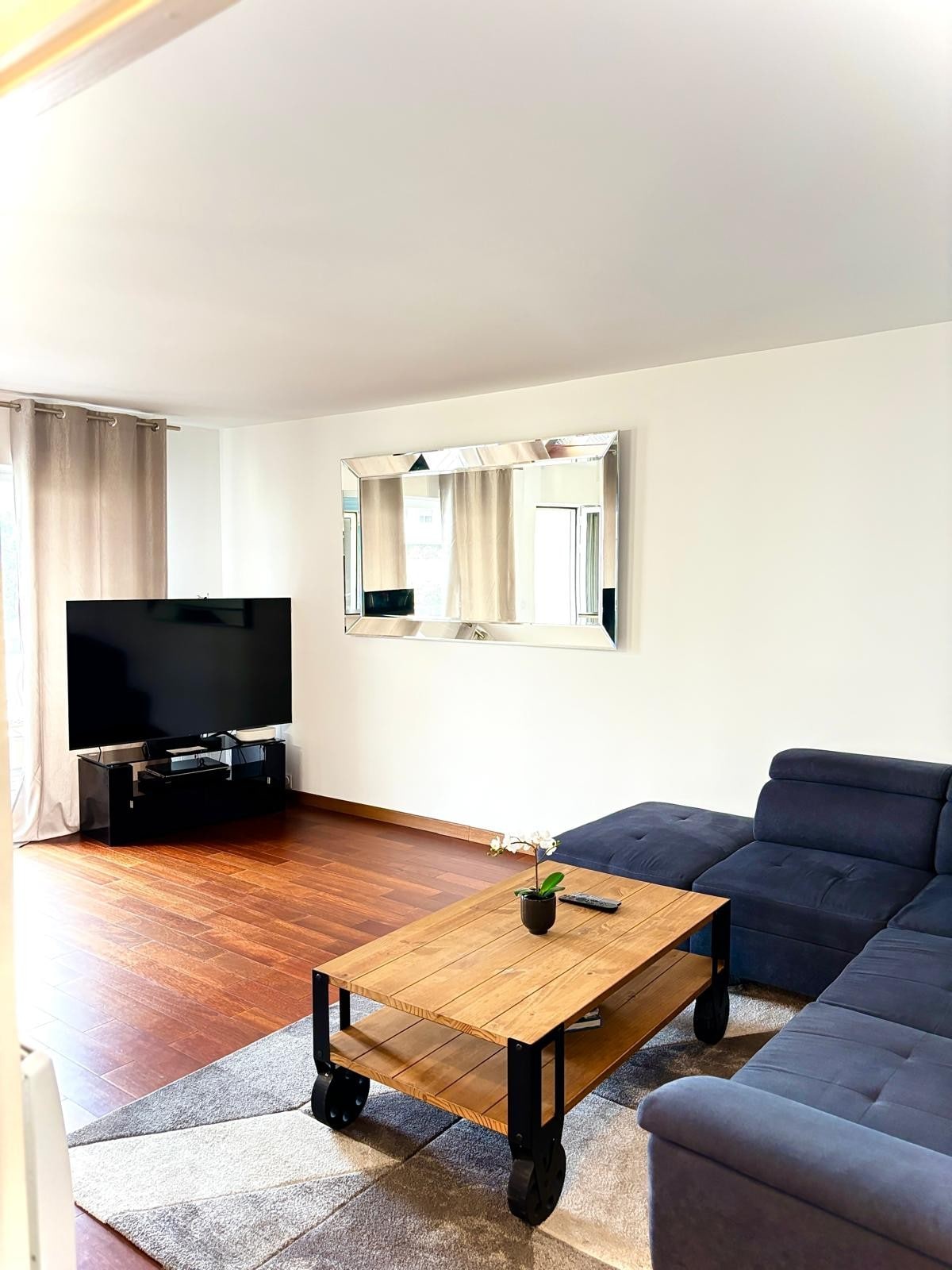 A living area features a modern sectional sofa and a wooden coffee table atop a soft area rug. A large television is positioned on a low stand, while natural light enters through the window, enhancing the space's brightness.