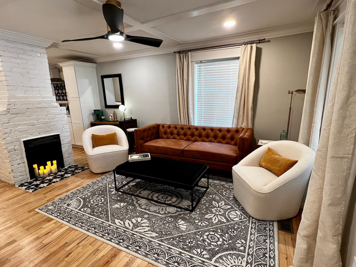 The living area features a stylish brown leather sofa complemented by two white accent chairs. A dark coffee table sits atop a decorative area rug, while a cozy fireplace is positioned nearby. Natural light enters through window treatments, enhancing the inviting atmosphere.