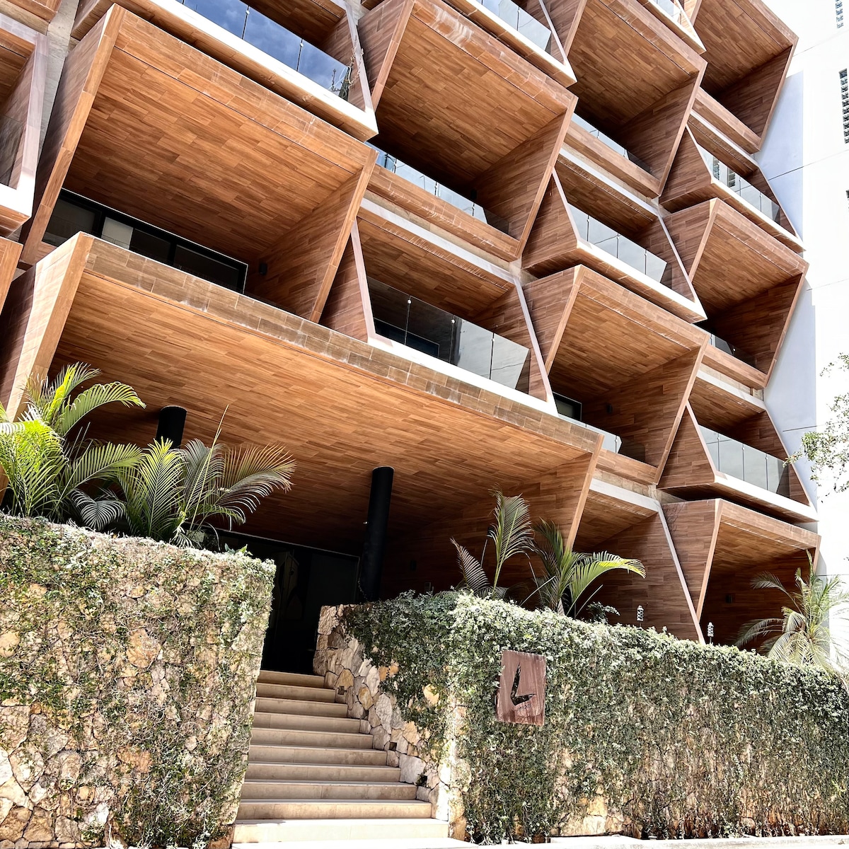 The building exterior features a modern design with distinctive geometric shapes and wooden facades. Lush greenery frames the entrance, which is accessed via a set of stone steps. A logo is visible on the stone wall, providing an inviting touch to the contemporary architecture.