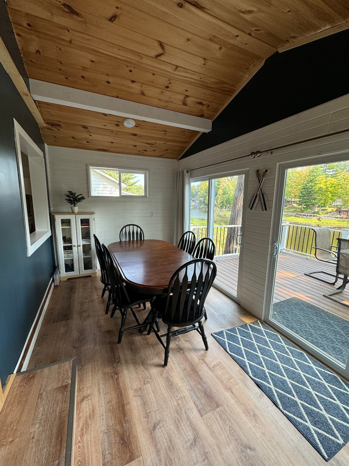 A dining area features a large wooden table surrounded by six black chairs. Natural light floods the space through two windows and a sliding glass door that leads to a deck, which offers views of the riverfront outside. Wooden beams accentuate the ceiling.