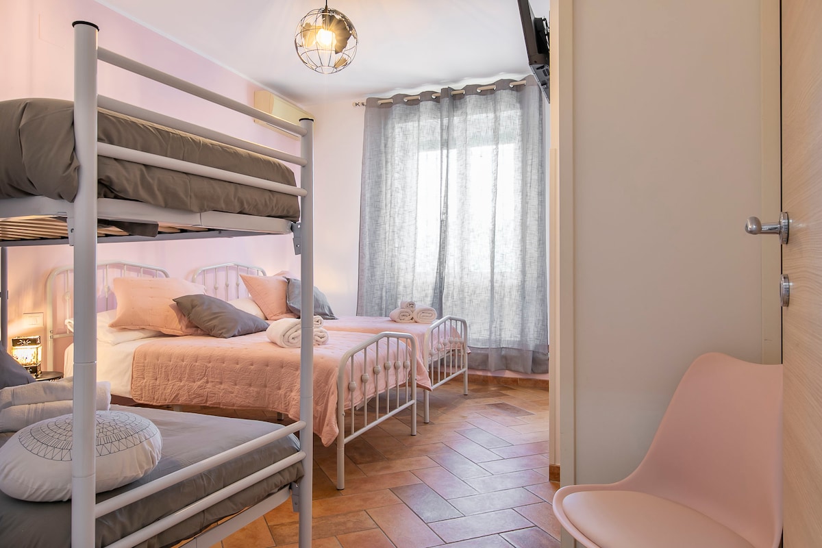 A well-lit bedroom features a combination of a bunk bed and a spacious single bed, with soft linens in neutral tones. Tall windows are dressed with sheer curtains, allowing natural light to fill the room. A modern chair is positioned nearby, adding a comfortable touch.