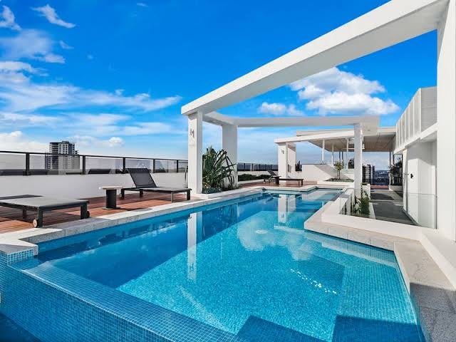 A rooftop pool area is depicted, featuring a clear blue water surface and modern lounge chairs positioned nearby. A shaded seating area is visible, alongside landscaped greenery. The space is bathed in natural light, with an unobstructed view of the sky enhancing the setting.