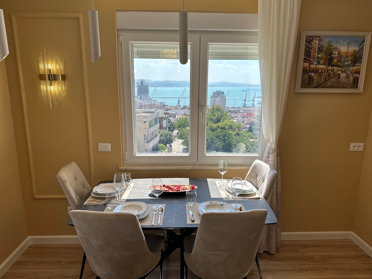 A dining table with four plush chairs is set by a large window, offering panoramic views of the sea and the cityscape. The table is elegantly arranged with tableware, and natural light fills the space, highlighting the warm hues of the walls.
