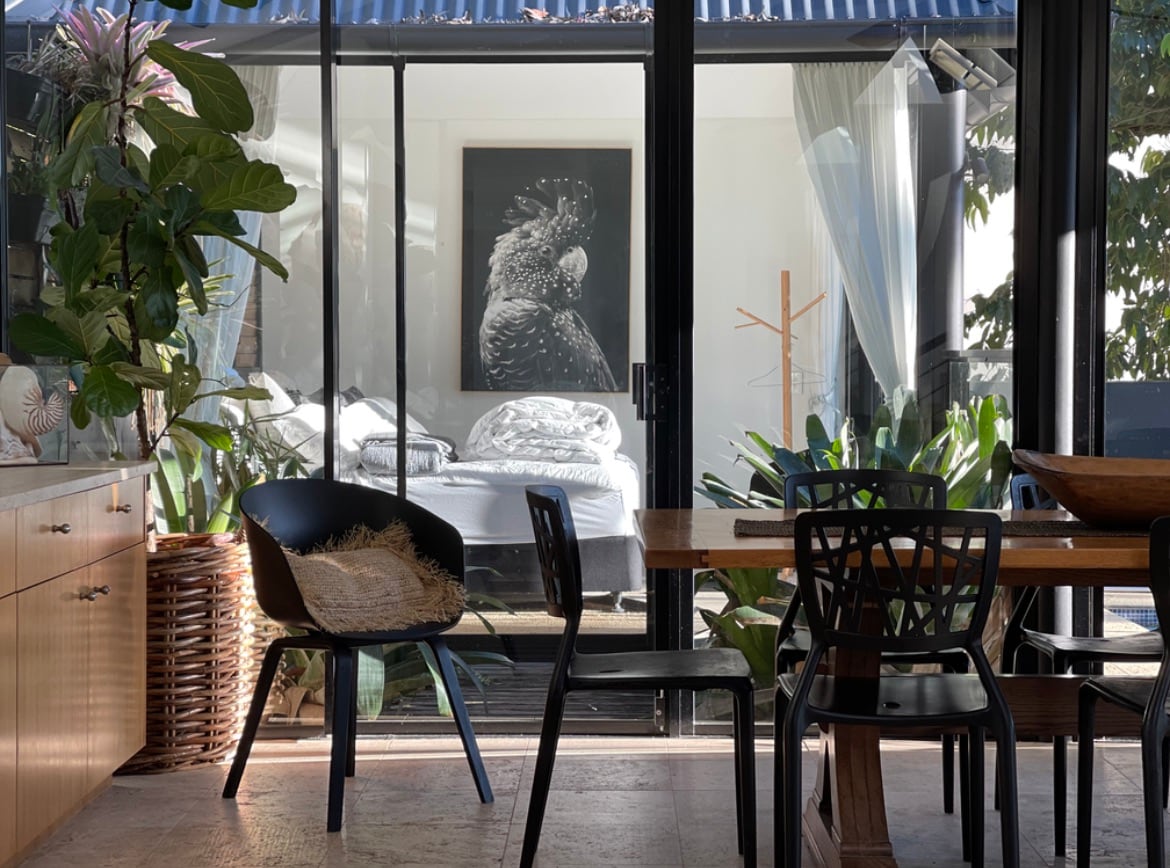 A dining area features a wooden table surrounded by black chairs, with a view through large glass doors into a light-filled bedroom. A striking artwork of a bird adorns the wall, while indoor plants add a touch of greenery to the inviting space.