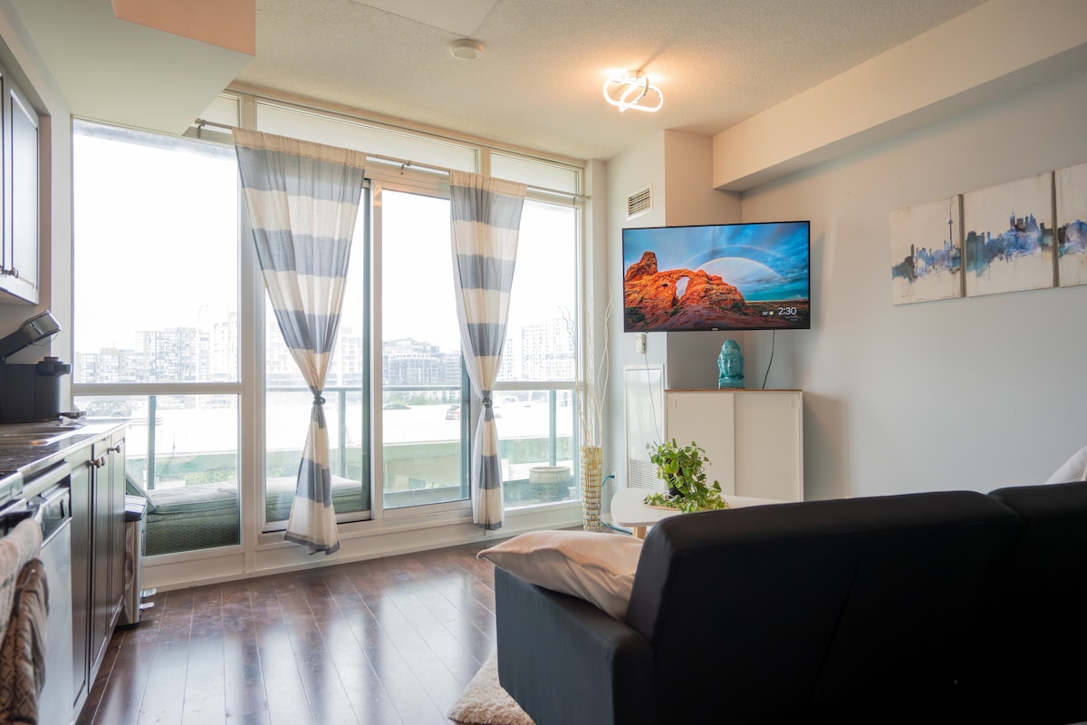 A spacious living area features large floor-to-ceiling windows with sheer striped curtains, allowing natural light to fill the room. A sleek black sofa faces a modern flat-screen TV, with a small plant adding a touch of greenery to the space.