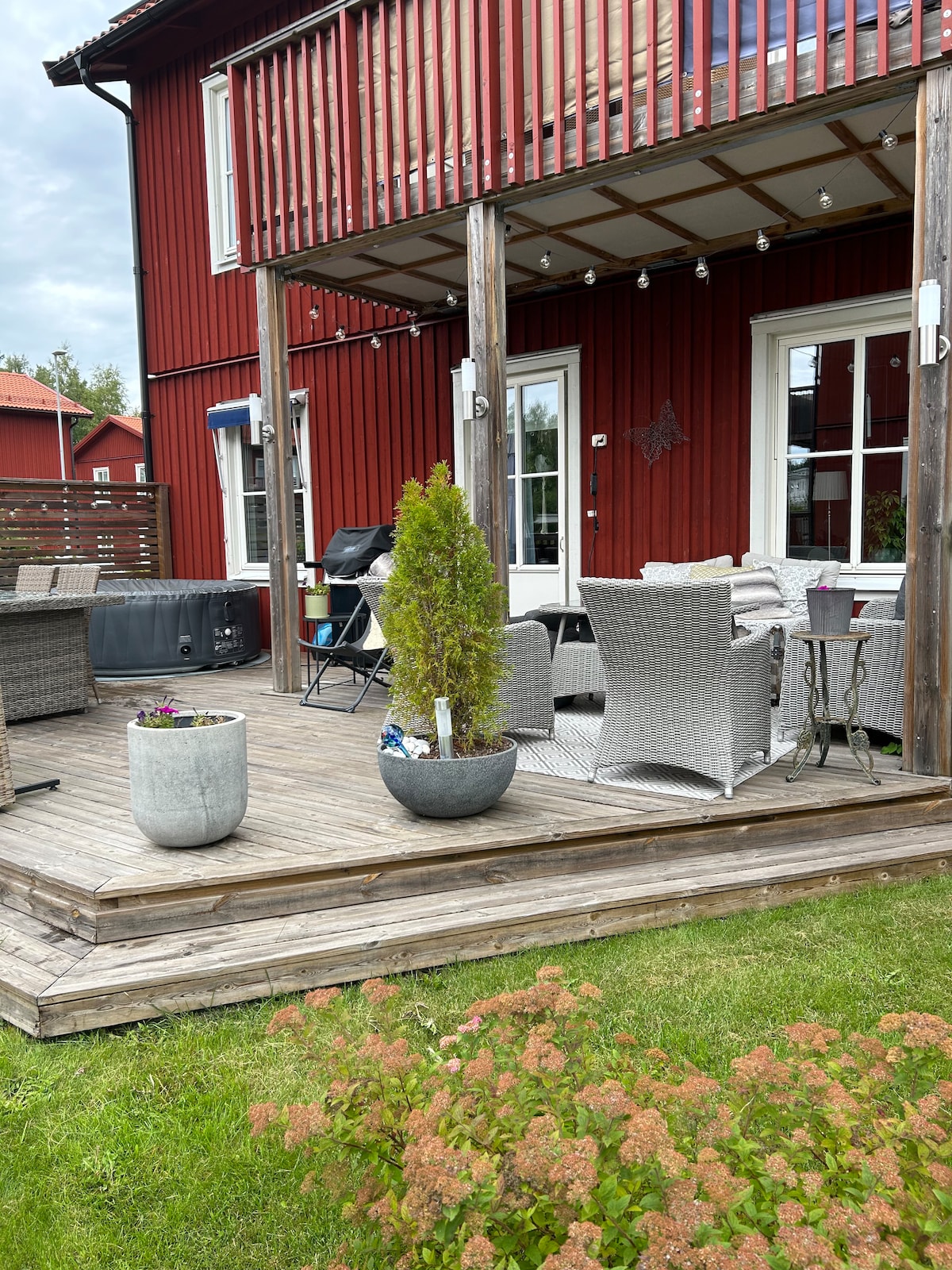 A spacious outdoor patio features comfortable seating arranged around a small table. Potted plants add greenery, while a barbecue grill is visible in the background. The wooden deck offers a clear view of the surrounding grass and landscaping.