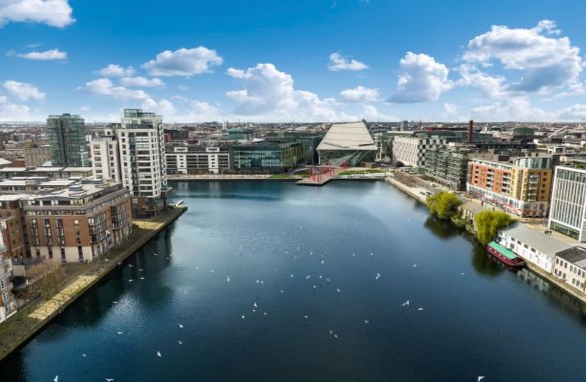 An aerial view captures the tranquil Grand Canal, flanked by modern buildings on both sides. The calm waters reflect the vibrant blue sky, dotted with fluffy clouds. The architectural features of the Bord Gais Energy Theatre and nearby cityscape create a harmonious urban scene.