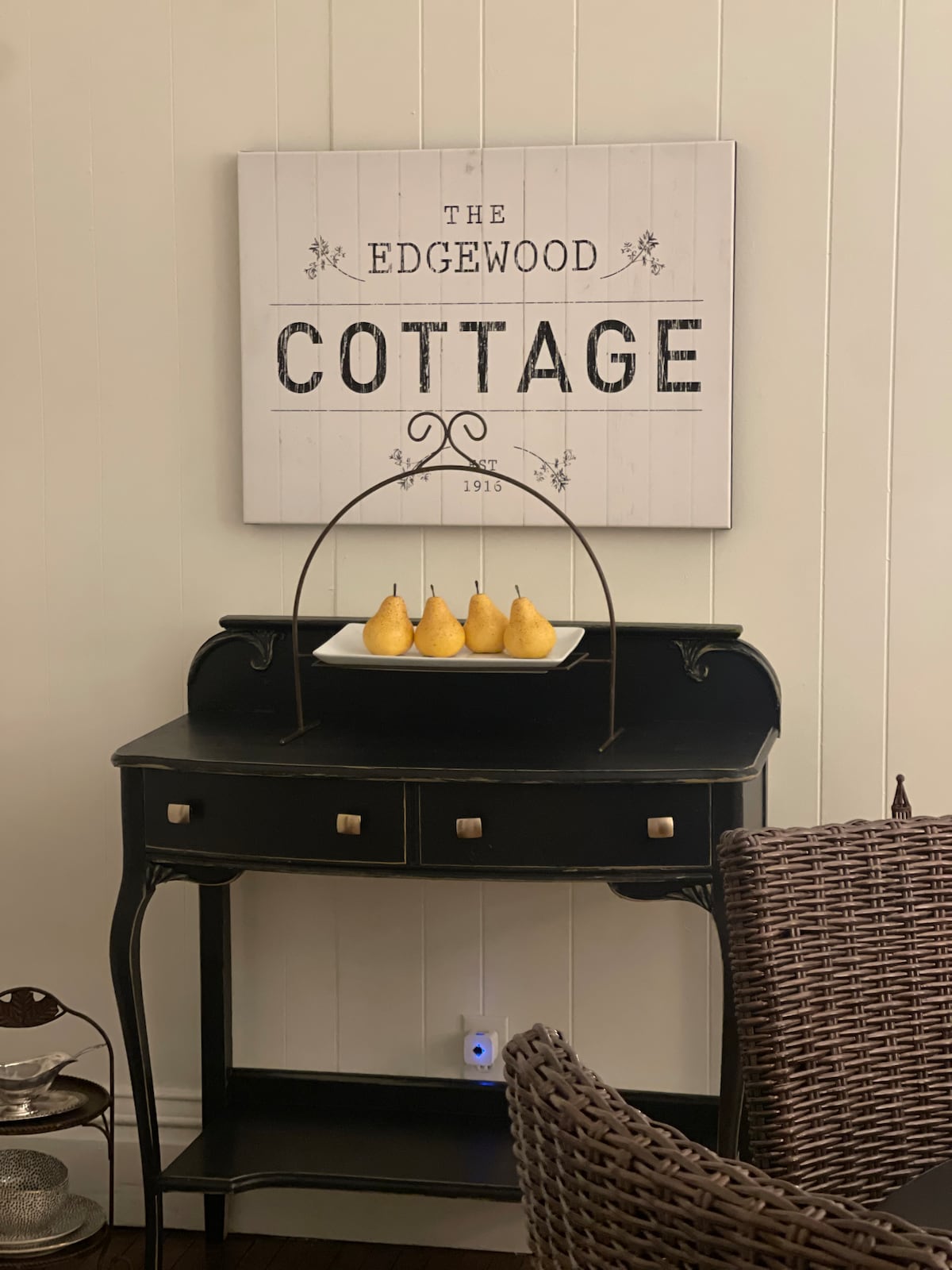A black side table is featured against a light wall. Above it hangs a sign that reads 'The Edgewood Cottage.' A decorative dish on the table holds several yellow pears, adding a touch of warmth to the space.