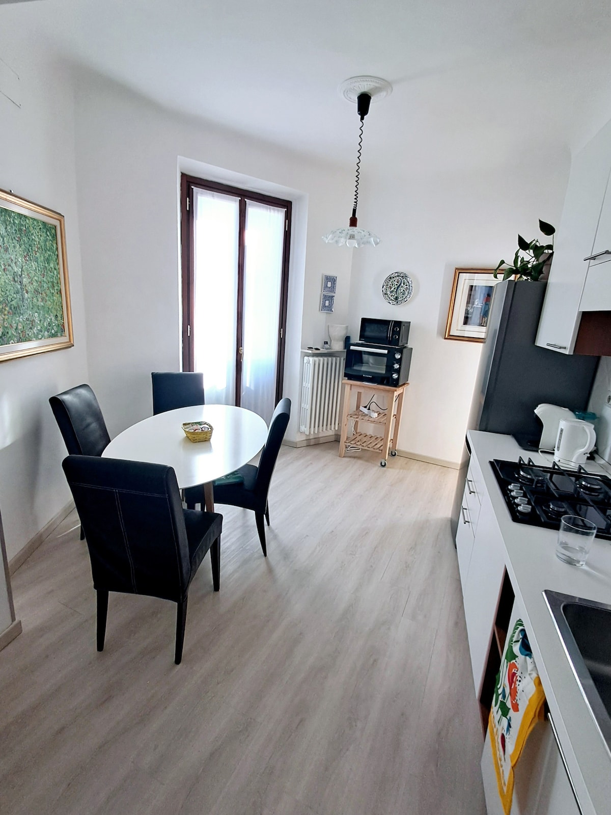 A bright kitchen area features a round white dining table surrounded by four black chairs. A radiator and a small cart with a microwave are present. Natural light enters through large windows with dark frames, illuminating the space and highlighting a wall adorned with a framed artwork.